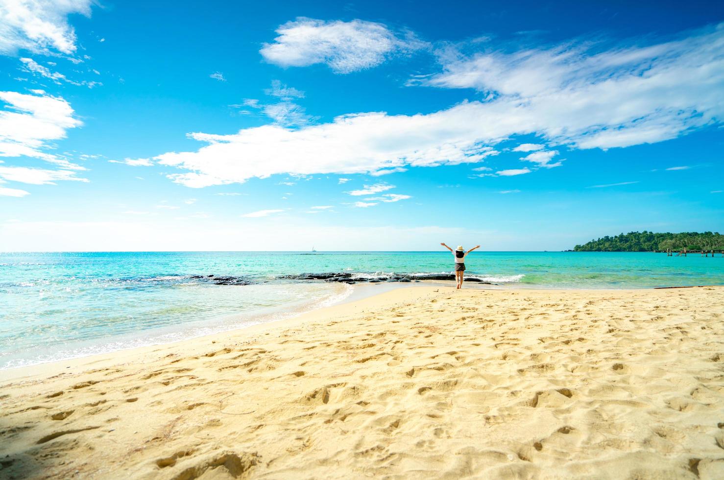 mujer joven feliz en camisas blancas y pantalones cortos caminando en la playa de arena. relajarse y disfrutar de unas vacaciones en la playa del paraíso tropical con cielo azul y nubes. chica en vacaciones de verano. vibras de verano. día feliz. foto