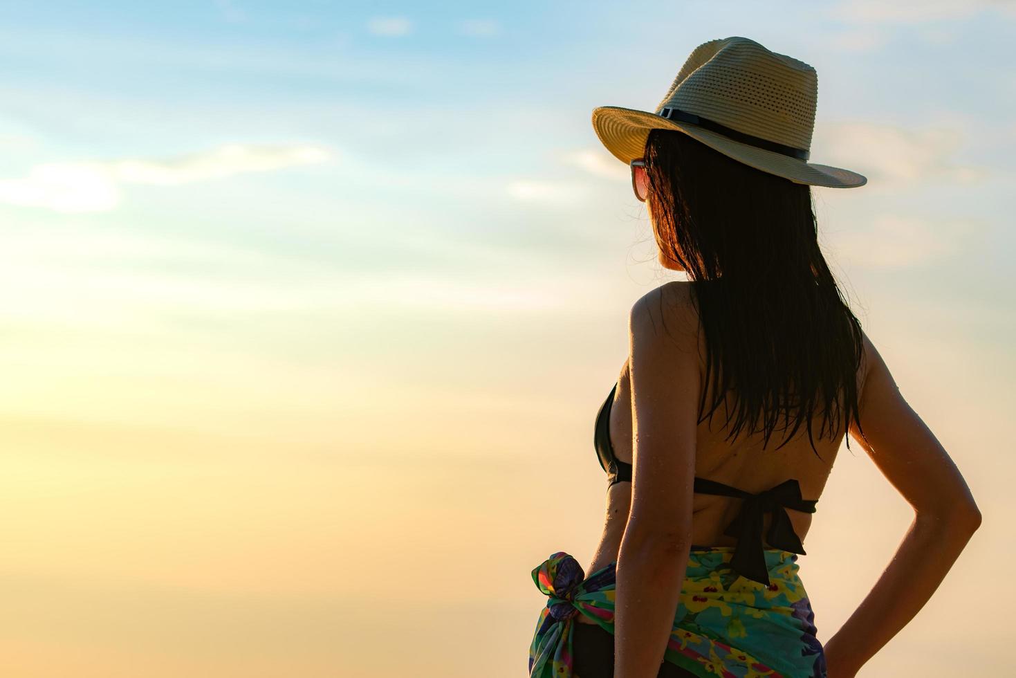 Back view of happy young Asian woman in black swimsuit and straw hat relax and enjoy holiday at tropical paradise beach at sunset. Girl in summer vacation fashion. Beauty sexy model. Elegant lifestyle photo