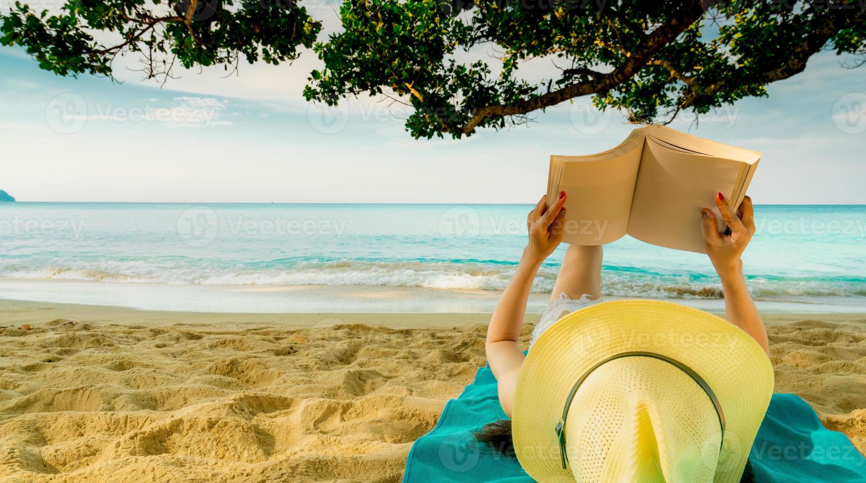 la mujer se acuesta en una toalla verde que se pone en la playa de arena debajo del árbol y lee un libro. vida lenta en vacaciones de verano. mujer asiática con sombrero relajándose y disfrutando de unas vacaciones en una playa tropical. vibras de verano foto