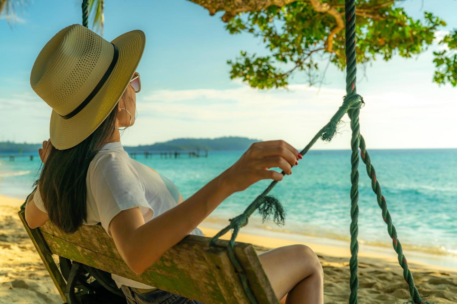 Young Asian woman sit and relax on swings at seaside on summer vacation. Summer vibes. Woman travel alone on holiday. Backpacker wear hat and sunglasses enjoy the day at tropical paradise beach. photo