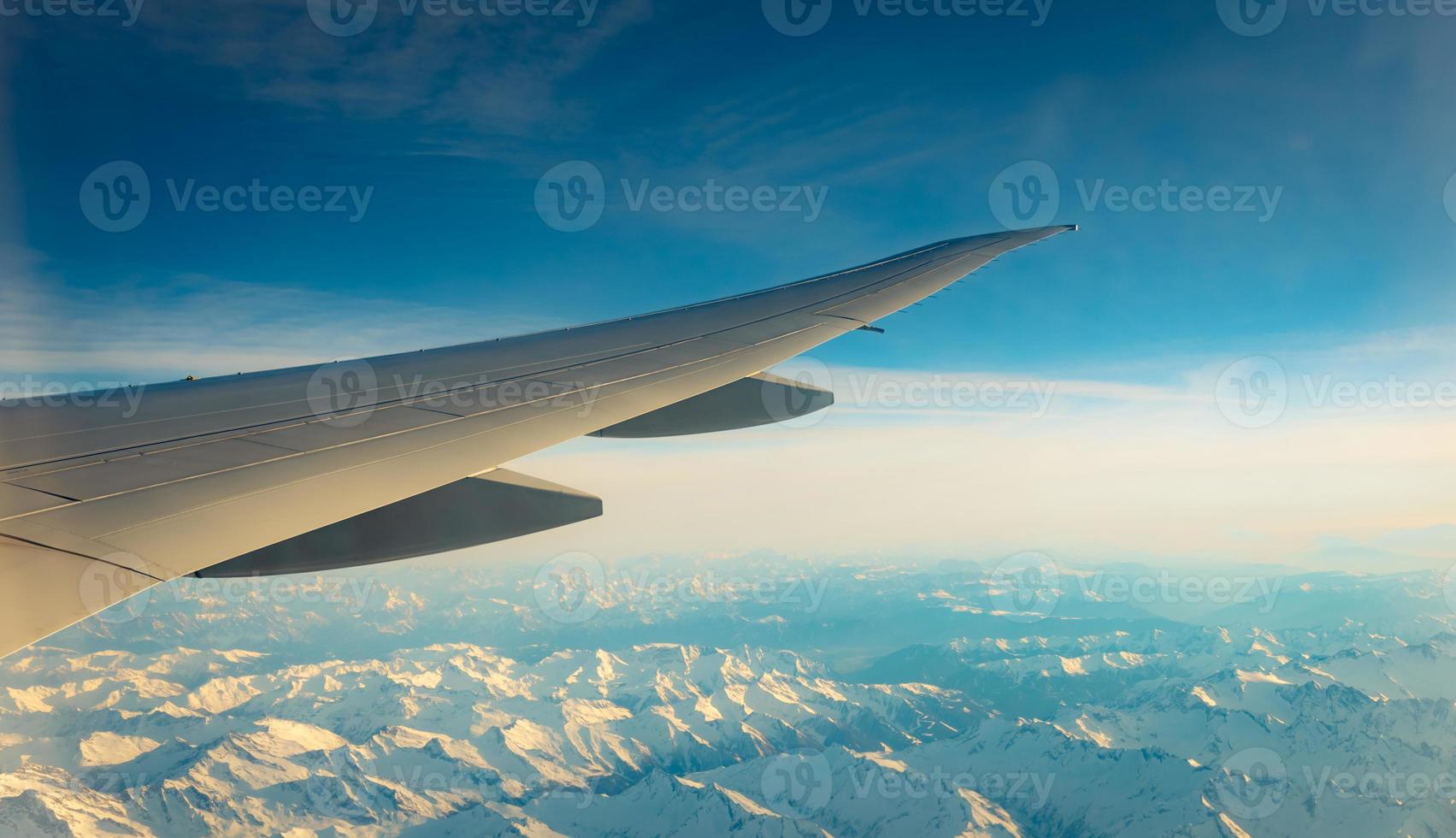 Wing of plane over mountain cover with white snow. Airplane flying on blue sky. Scenic view from airplane window. Commercial airline flight. Plane wing. Flight mechanics concept. International flight. photo