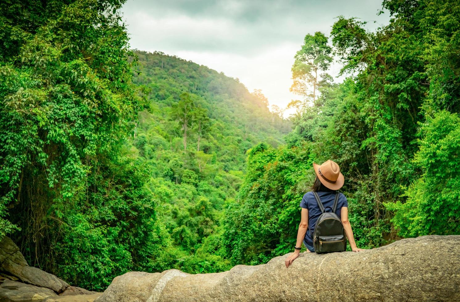 mujer feliz viaja sola en el bosque. mujer activa con buen humor sentada en piedra en un valle verde con un árbol denso en el bosque. Felices vacaciones. vista posterior de una mujer feliz con mochila, banda inteligente y sombrero. foto