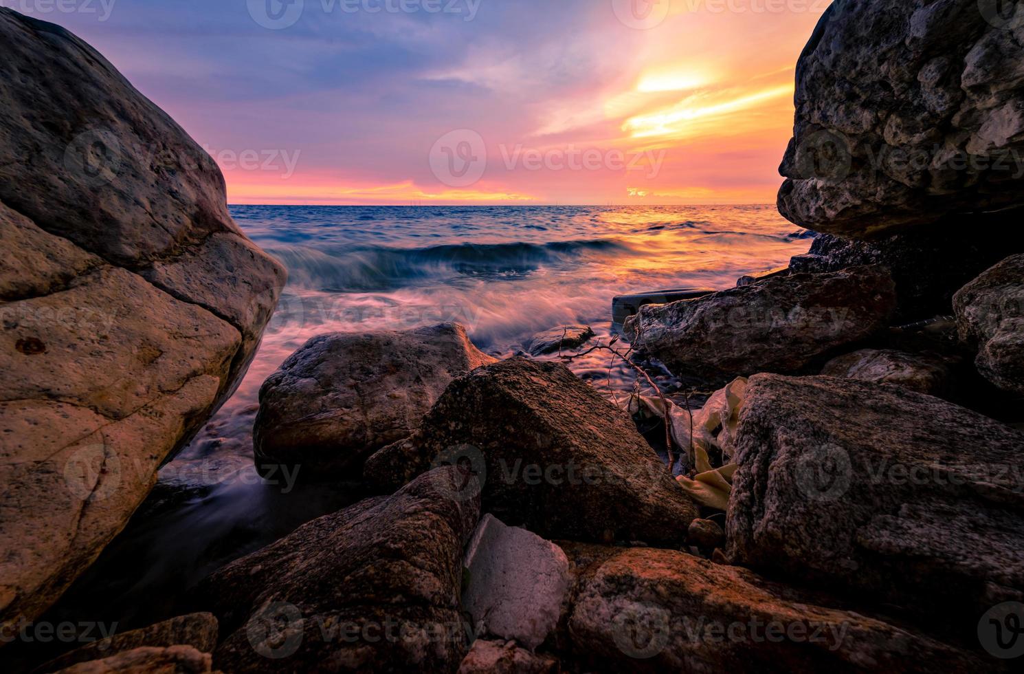 Ocean water splash on rock beach with pink and golden sunset sky. Sea wave splashing on stone at sea shore on summer. Tires and plastic sacks on tropical beach. Beach and ocean pollution. Soft wave. photo