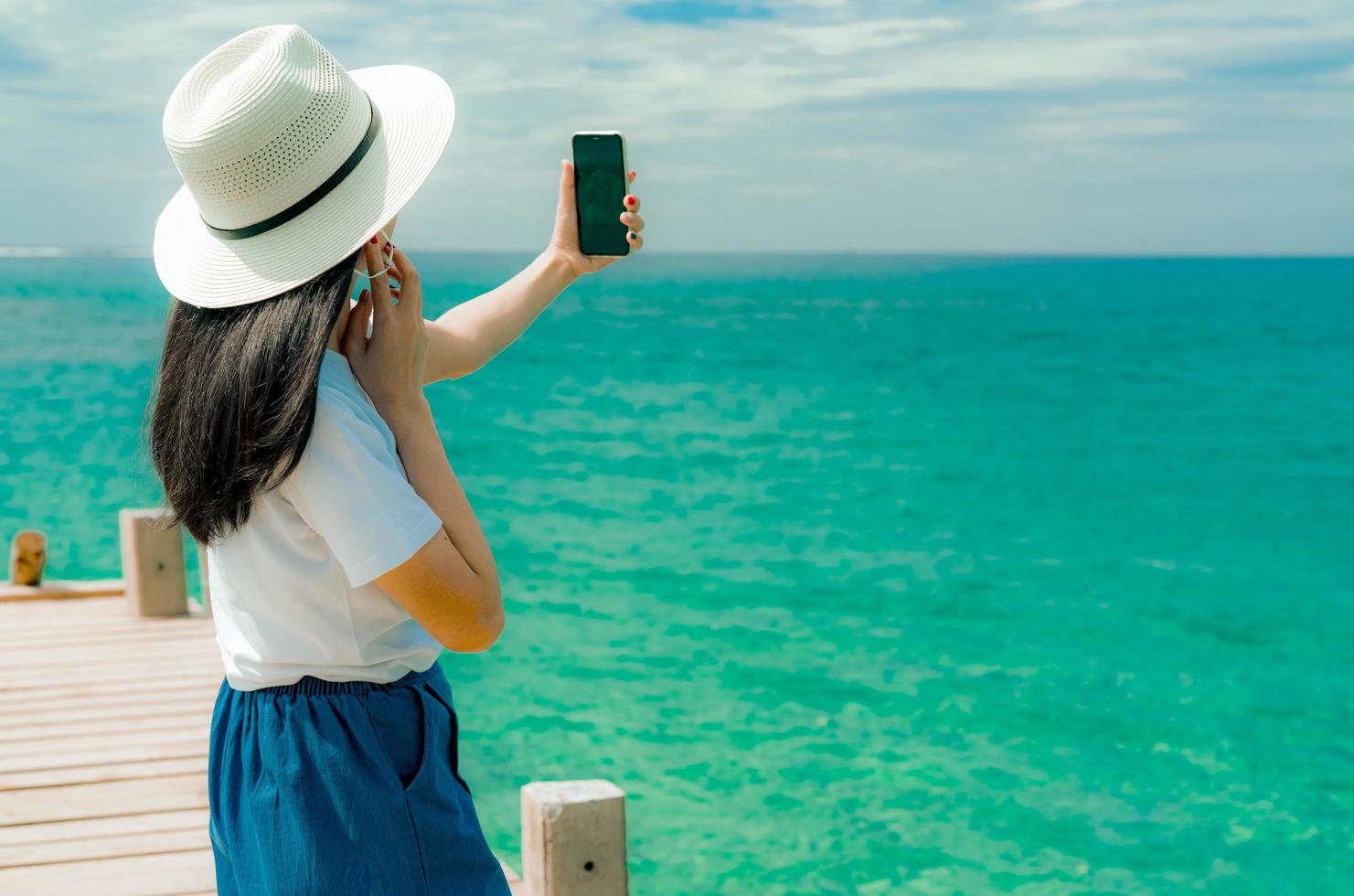 Young Asian woman wear hat in casual style use smartphone taking selfie at pier. Summer vacation at tropical paradise beach. Happy girl travel on holiday. Woman enjoy and relax life. Summer vibes. photo