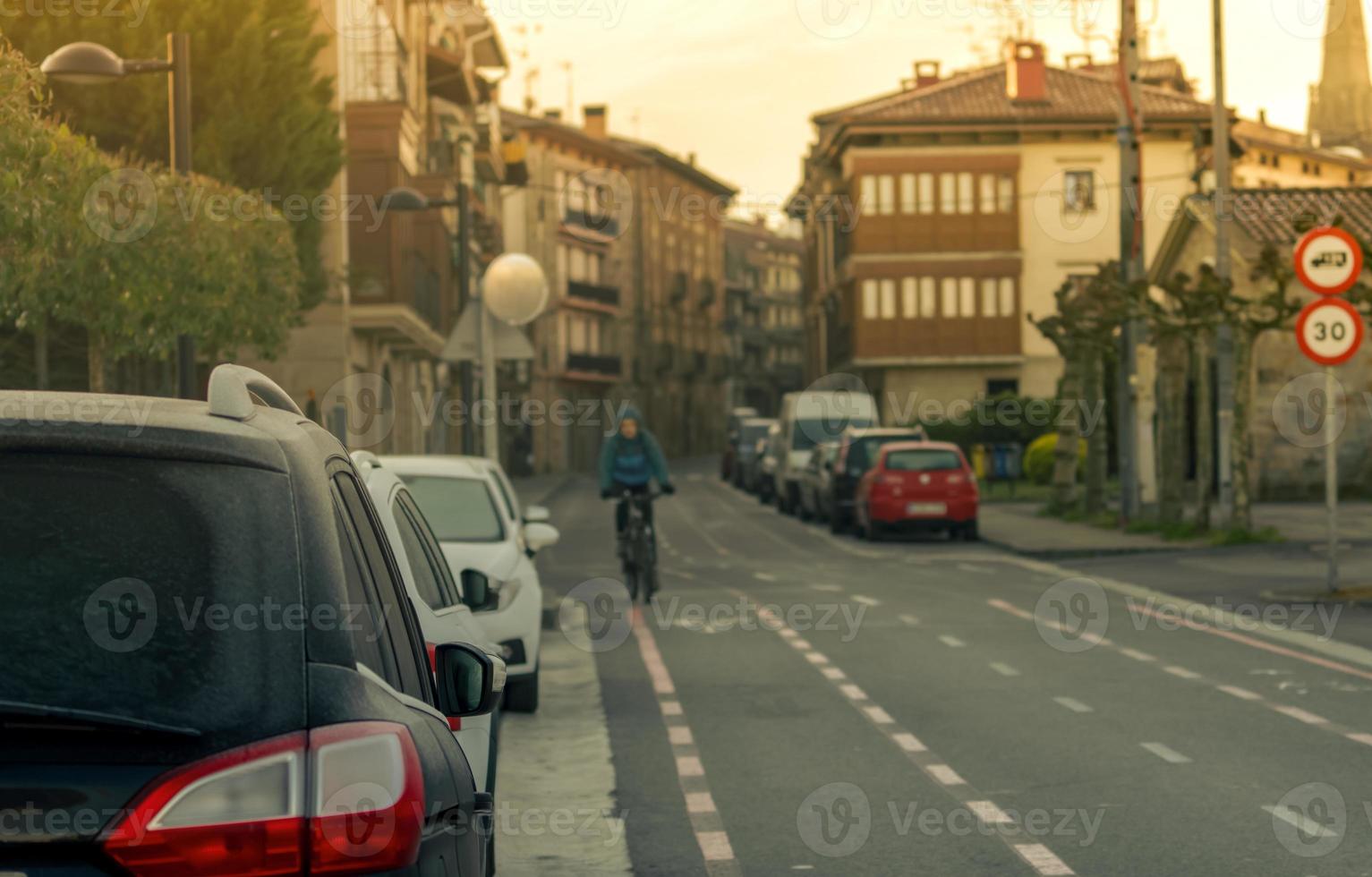 Rear view of car parked beside asphalt street in town with blurred people ride a bicycle for exercise in spring morning. Blurred traffic sign of city limit speed 30 km hr. Old building in the city. photo