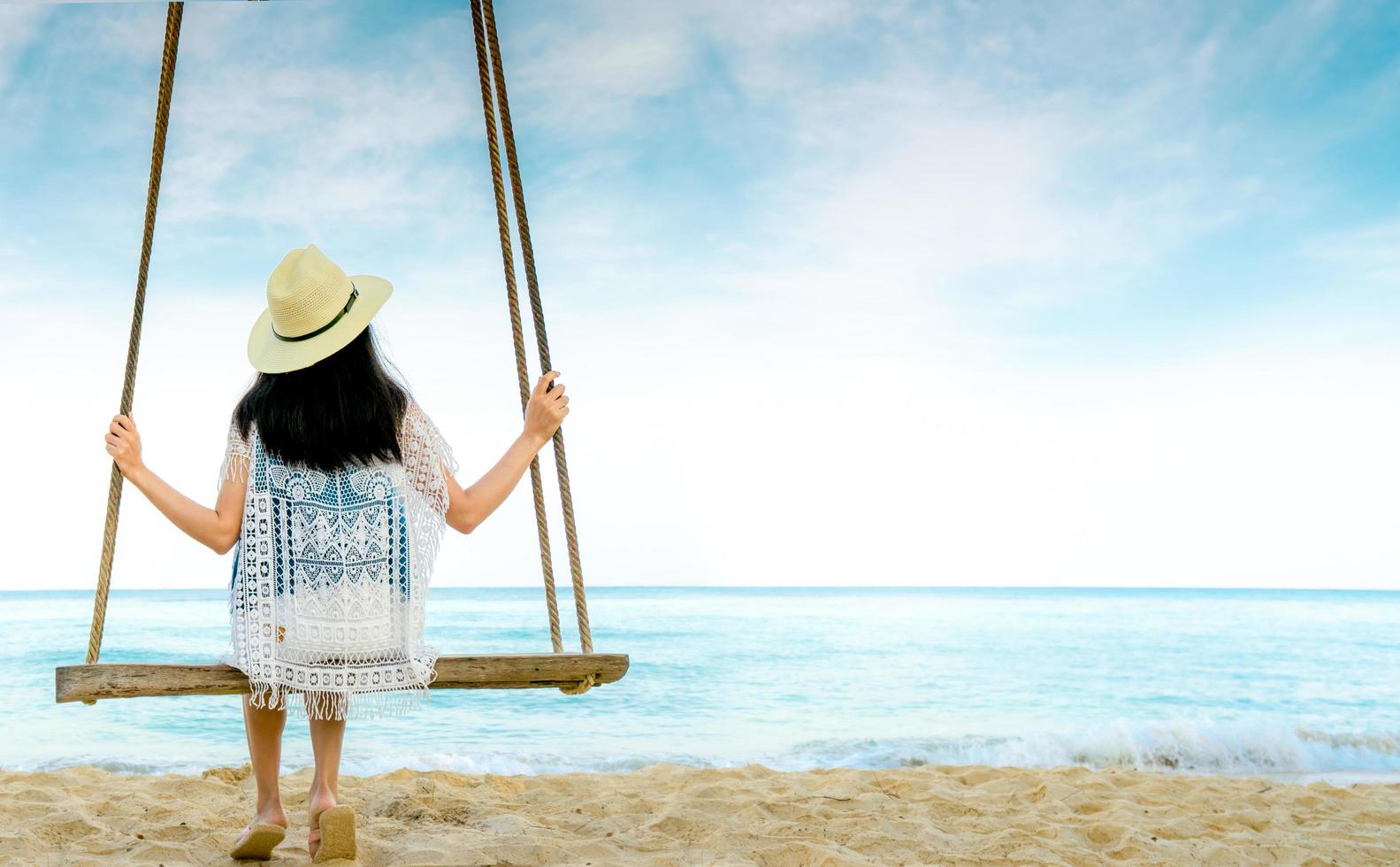 Asian woman in casual style wear hat and sandals sit on swings at sand beach  and looking beautiful tropical paradise sea and sky. Summer vacation. Summer vibes. Enjoying and relaxing girl on holiday. photo
