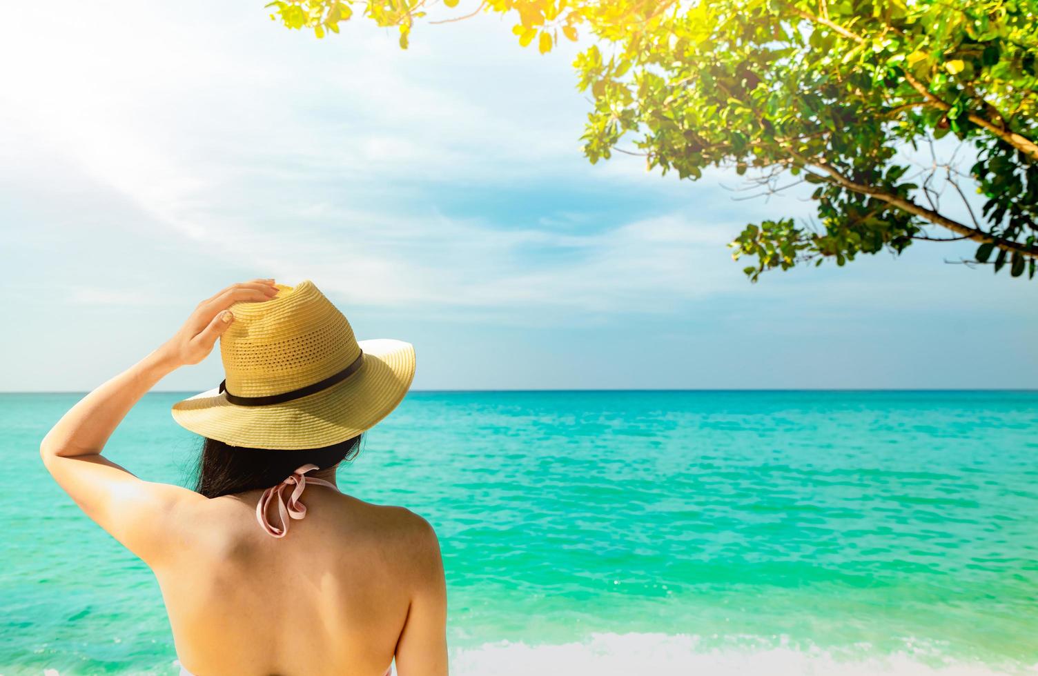 Back view of sexy and happy young Asian woman wear straw hat relaxing and enjoy holiday at tropical paradise beach. Girl standing at the beach and looking the sea in summer vacation. Summer vibes. photo