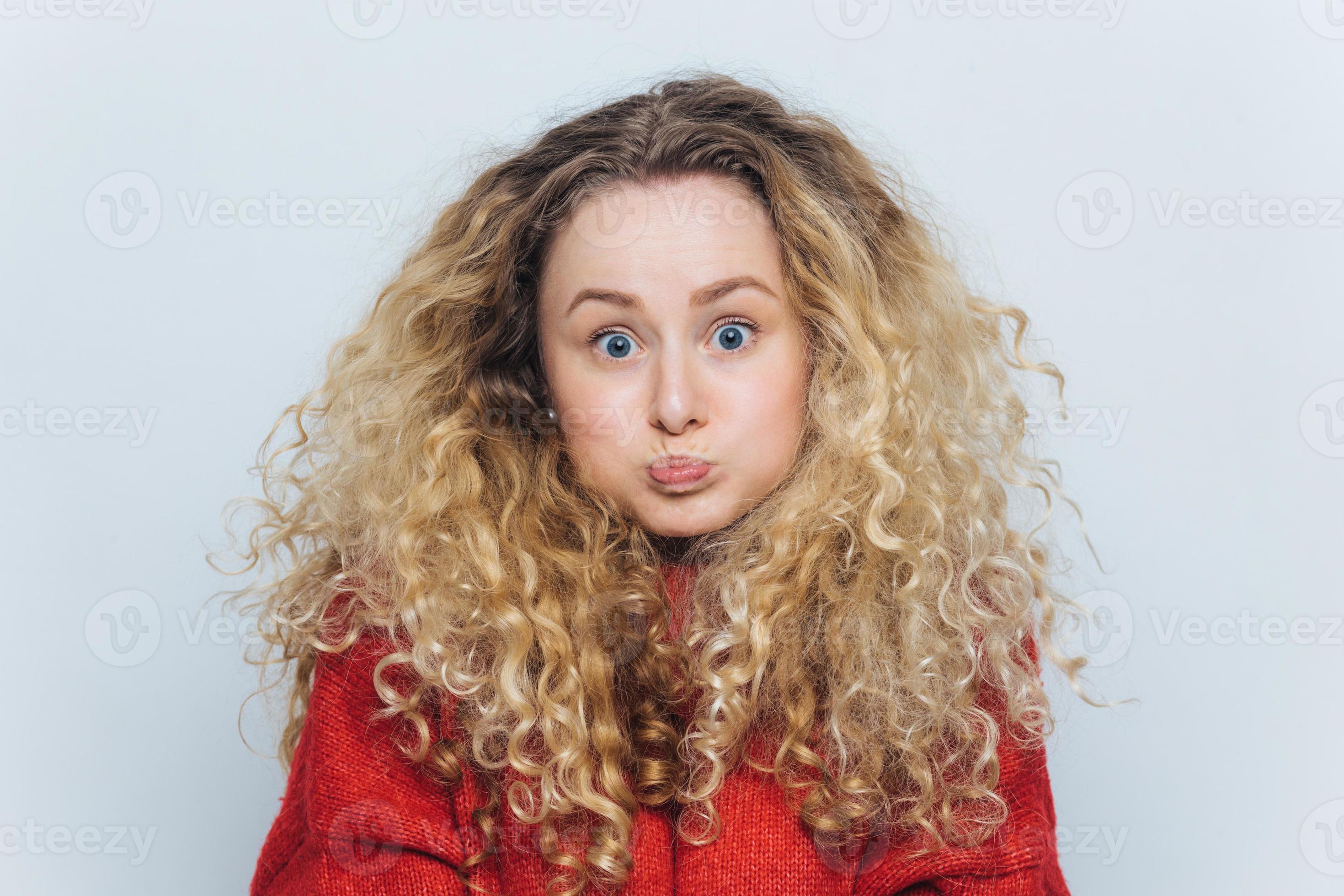 Fuuny Blonde Female With Curly Hair Blows Cheeks And Stares At Camera Reacts Actively On Unexpected News Isolated Over White Background Emotional Young Woman Poses Indoor Body Language Concept Stock Photo