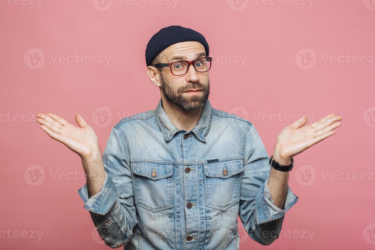 Indoor shot of hesitant bearded man shrugs shoulders, looks uncertain, doubts when makes decision, isolated over pink background. People, doubt and hesitation concept. Body language concept. photo