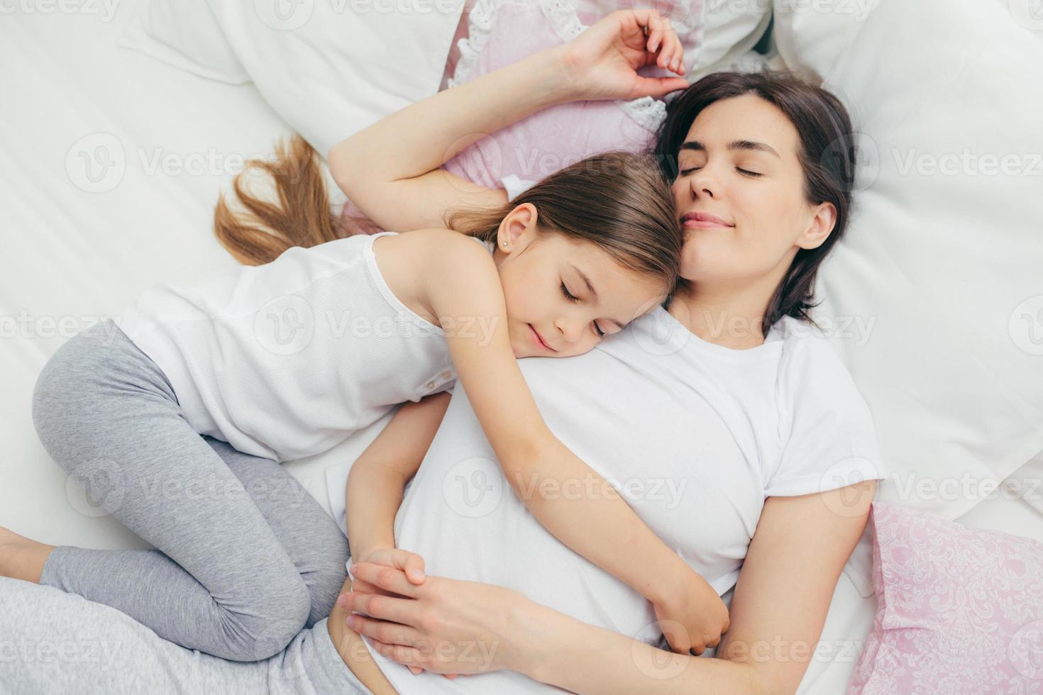 Pleased little child sleeps near her mother, embraces with love, has pleasant dreams, lie on comfortable bed. Mum and cute daughter have good sleep in bedroom. Family, sleeping and rest concept photo