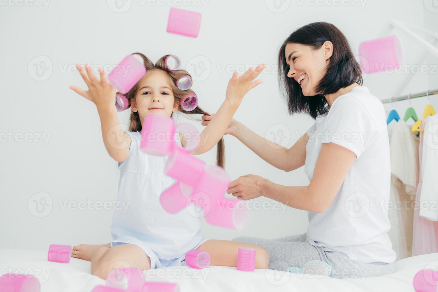 Funny female child throws curlers in air, has fun together, her mother does hairstyle, wear casual clothes. Beautiful woman winds curlers on hair, sit in bedroom. People, family and motherhood concept 7671396 Funny female child throws curlers in air, has fun together, her mother does hairstyle, wear casual clothes. Beautiful woman winds curlers on hair, sit in bedroom. People, family and motherhood concept 7671396