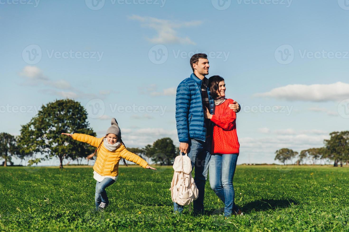 Young couple in love stand on green grass, embrace and look pensively. Playful small child enjoys freedom, runs on field, plays near her parents, has good mood and recreation. Peope and nature photo