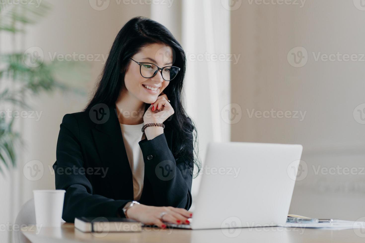 Cheerful female employee types on laptop computer, works remotely, concentrated with smile at device screen, writes email letter, works in office drinks coffee uses new application. Technology concept photo