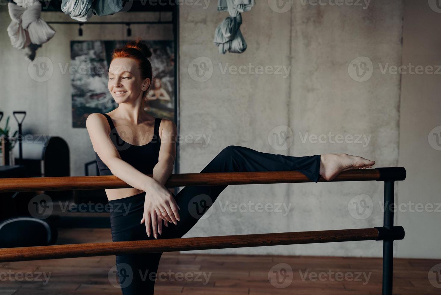 Slender smiling red haired woman stretching leg on ballet barre in classroom photo