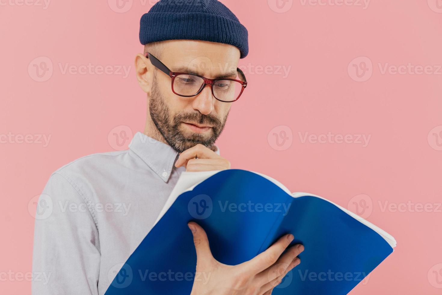 Close up shot of serious man holds chin, has dark stubble, wears glasses for good vision, carries opened book, learns information, stands against pink background, involved in reading, has clever look photo