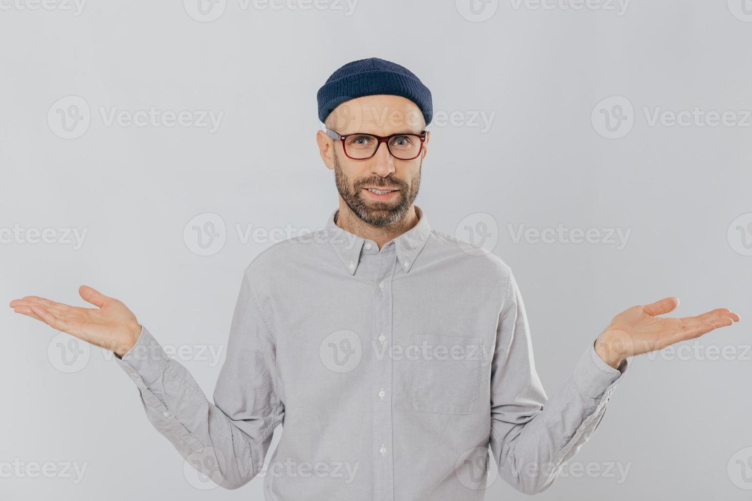 Photo of handsome man with uncertain expression, spreads hands, wears glasses and headgear, isolated over white backgrounnd. Unshaven young Caucasian male shruggs shoulders in bewilderment indoor