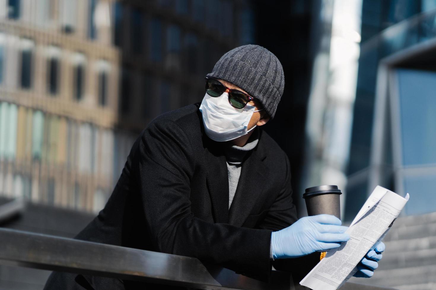 joven en la calle con café para llevar y periódico, usa máscara y guantes médicos protectores, evita la propagación del coronavirus, mira a un lado pensativamente, posa al aire libre. pandemia de covid-19, cuarentena foto