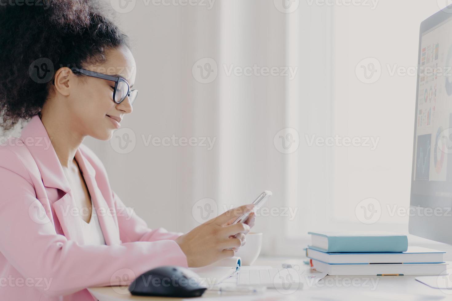 Horizontal shot of focused curly haired female office worker holds mobile phone, searches information in internet, wears optical glasses and elegant suit, computer monitor in front, does paper work photo