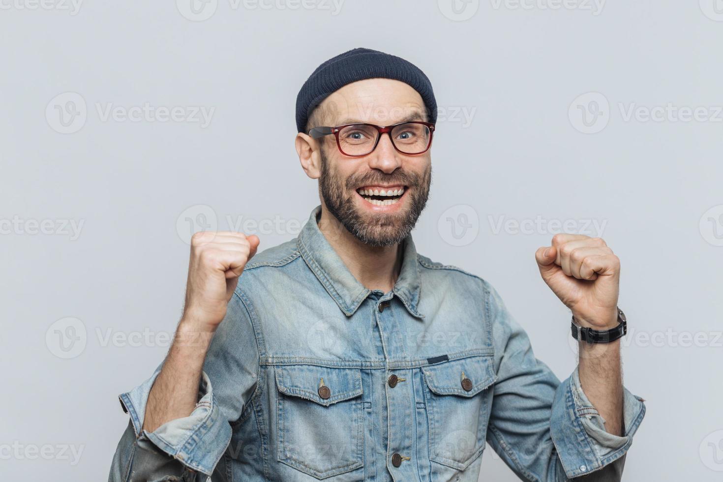 Portrait of happy successful man rejoices his triumph, clenches teeth, has overjoyed expression, isolated over grey background. Handsome fashionable male celebrates his victory. Achievement concept photo