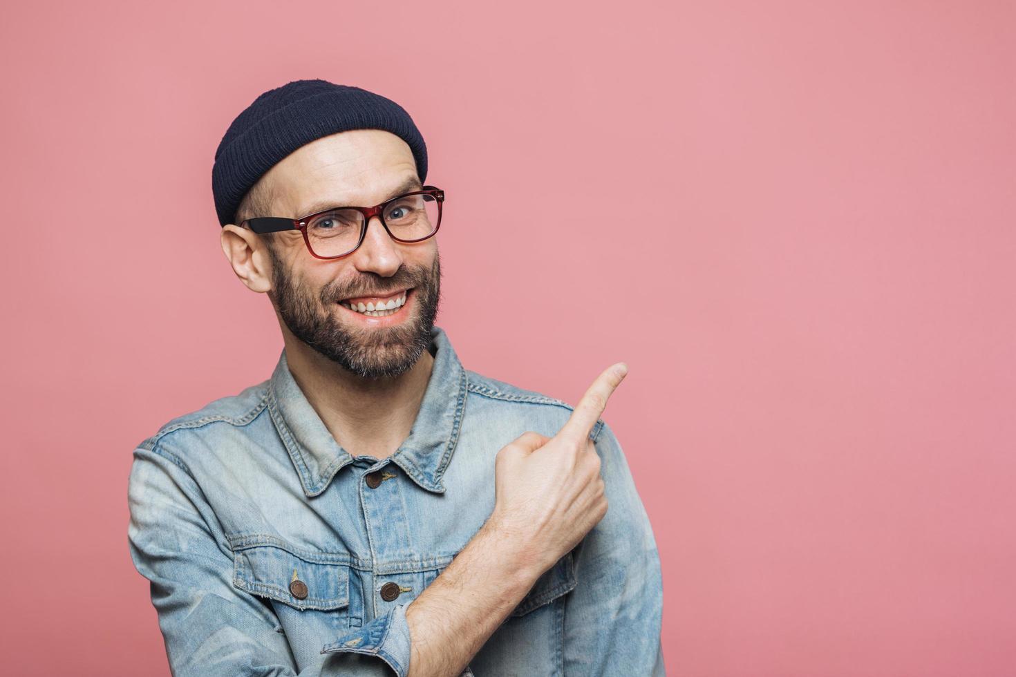 Advertisement concept. Cheerful young bearded man with glad expression wears eyewear, denim jacket and hat, indicates at copy space for your advertisment or promotional text, has happy look. photo