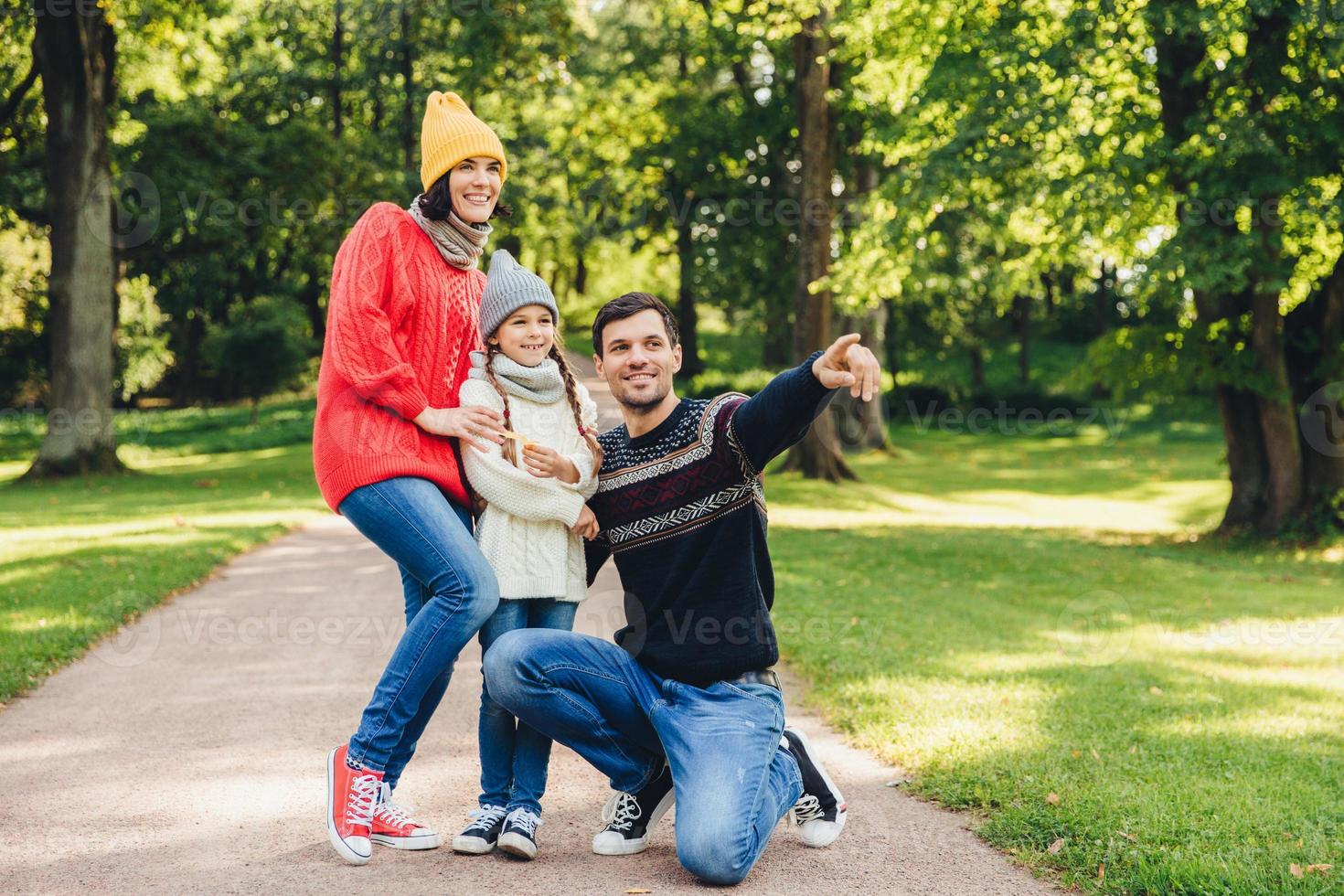 Horizontal portrait of family with good relaionship stand close to each other, admire landscapes or beutiful nature. Happy father shows to her daughter big old tree in park, indicates with fore finger photo