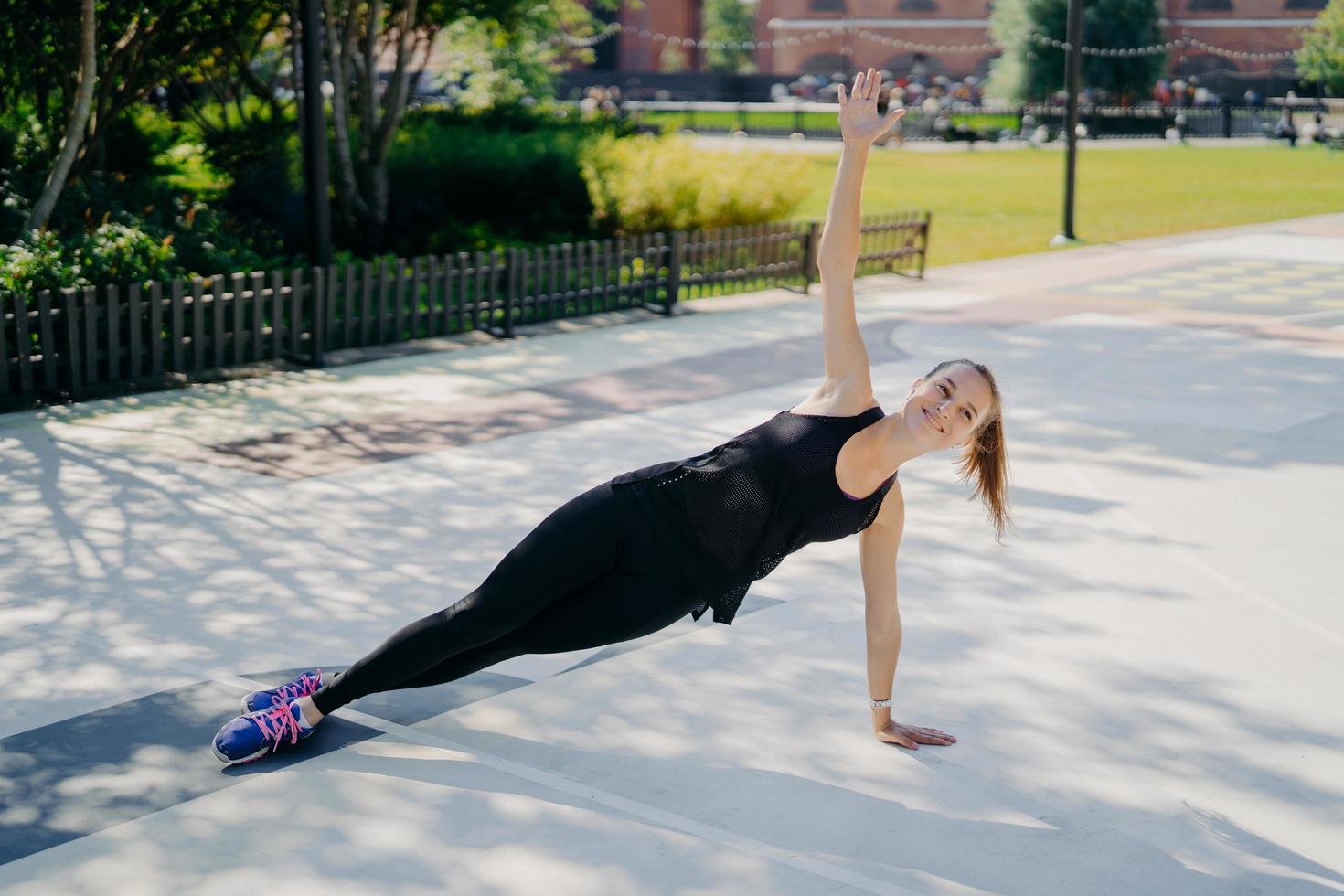 Sport and recreation concept. Athletic slim woman stands in sideways plank raises arm smiles positively dressed in active wear leads healthy lifestyle strengthens core muscles improves stability photo