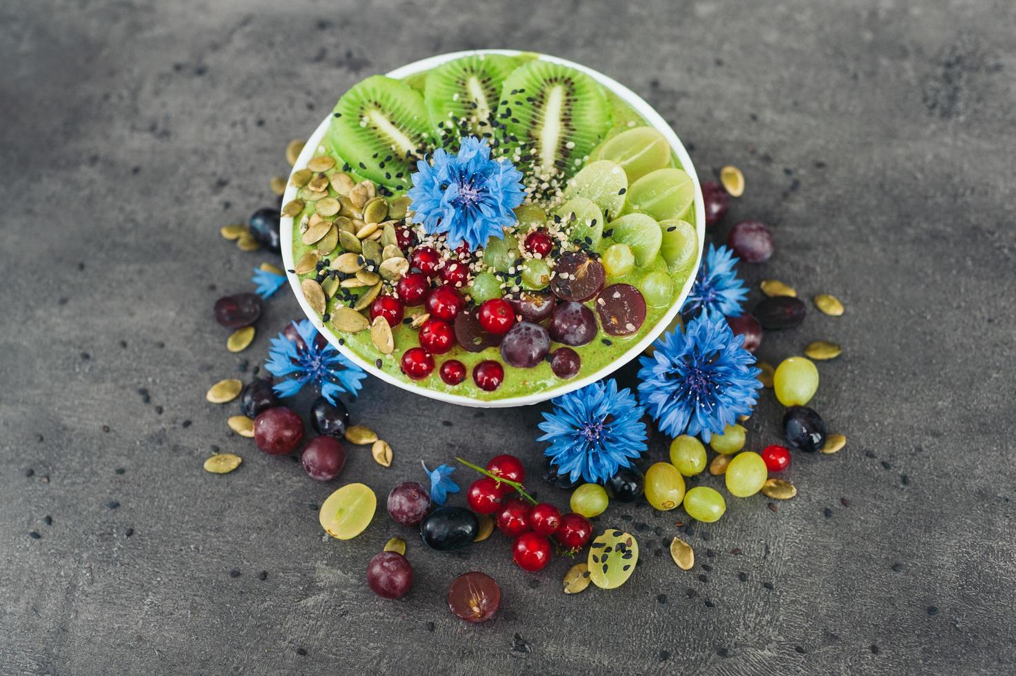 Bowl with fresh kiwi salad, seeds, red current, green grape and blue cornflowers around. Vegeterian dish of ripe fruit. Top view. Blank space photo