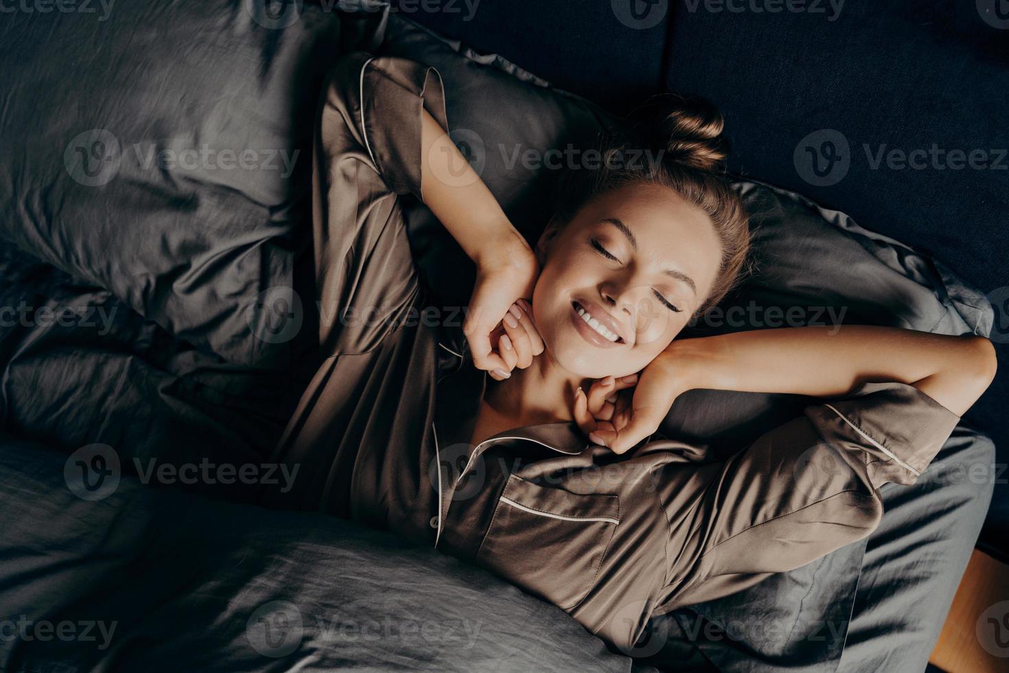 Young beautiful relaxed brunette female in satin pajama stretching in bed photo