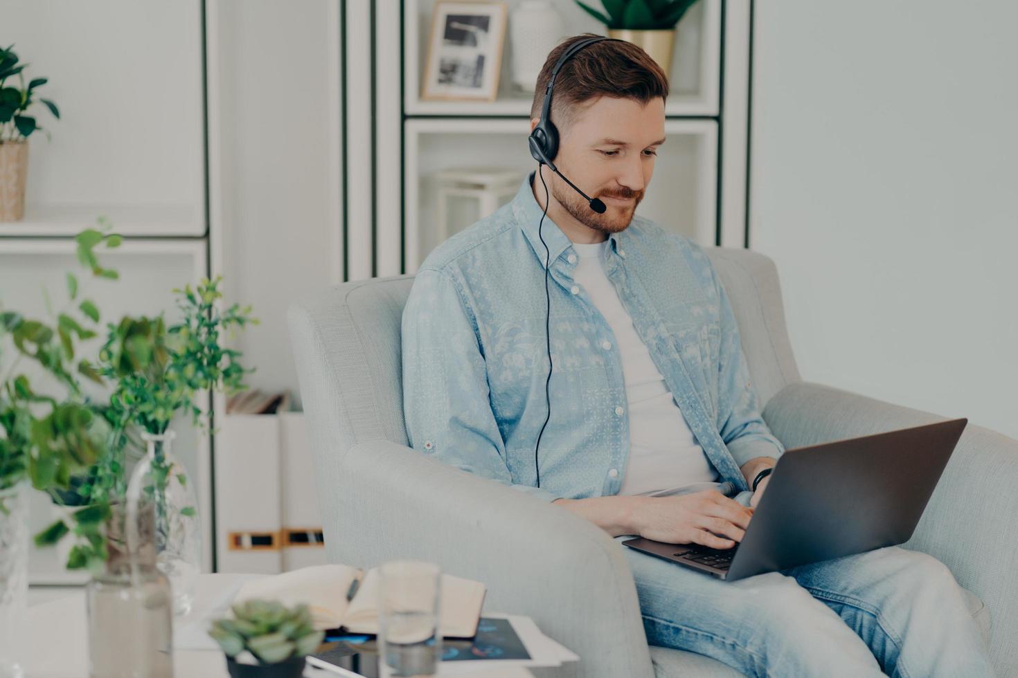 Busy man dressed in denim clothes supplies consultant service types on laptop computer wears headphones with microphone works distantly sits in comfortable armchair participates in online negotiations photo