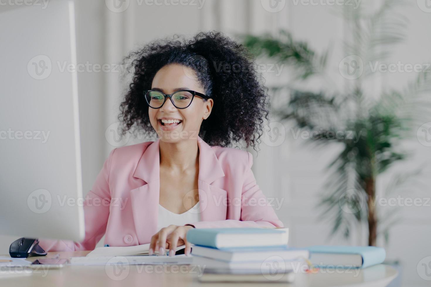 Horizontal shot of cheerful African American freelancer browses internet on computer, focused at screen, wears optical glasses and rosy formal costume, poses at desktop with pile of books, papers photo