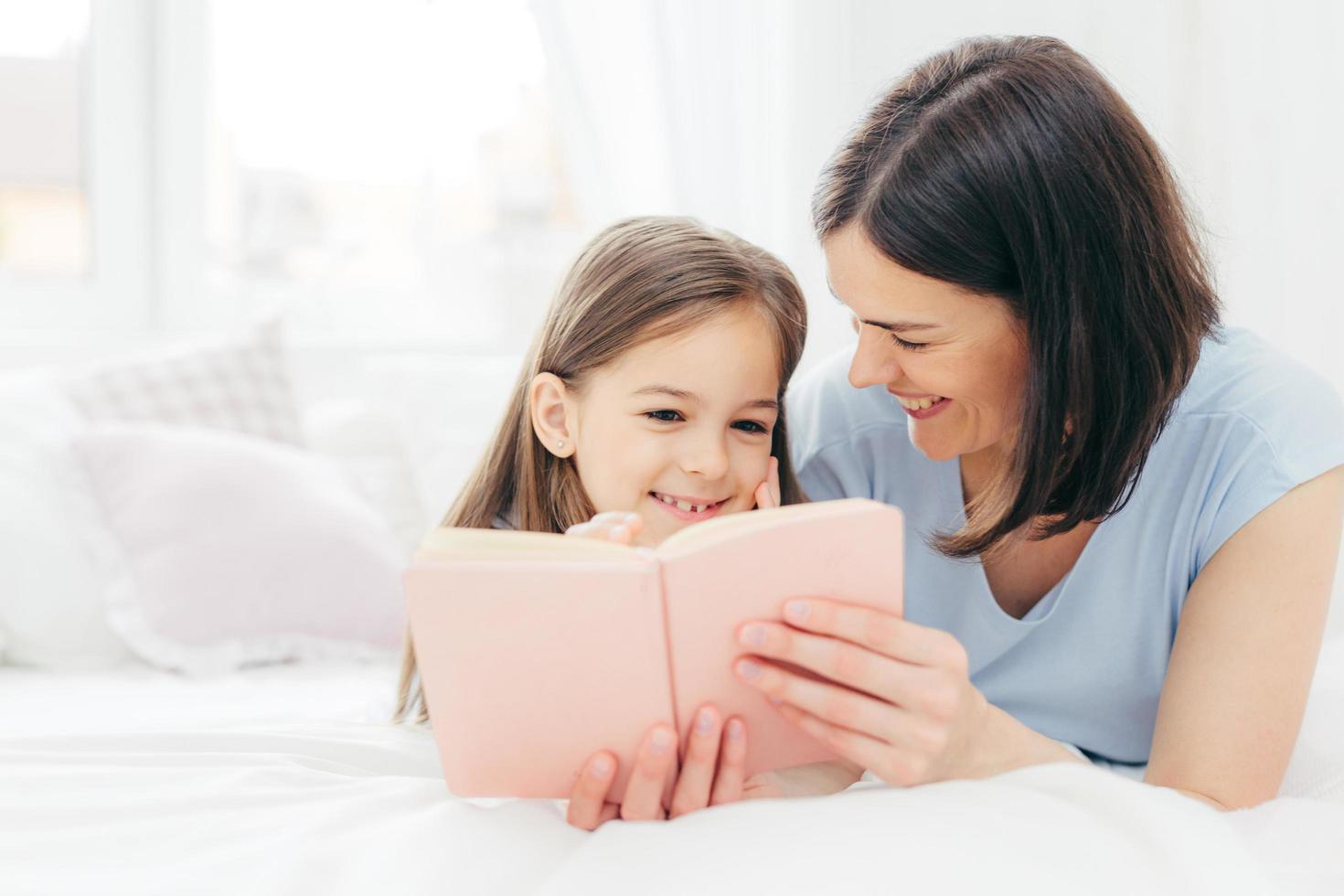 toma interior de una niña de aspecto agradable con expresión curiosa, lee un libro interesante junto con su afectuosa madre, ve imágenes coloridas, se acuesta en la cama en un amplio dormitorio. infancia foto