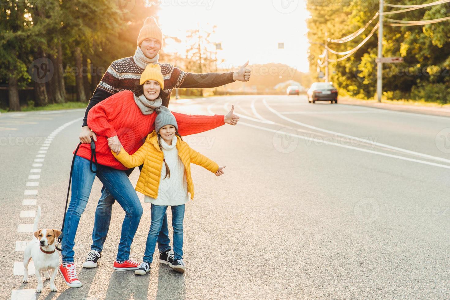 Portrait of family mother, father and little daughter walk with pet, stand on road, raise thumbs as show ok sign, express approval or agreement, have good mood after spending time on fresh air photo