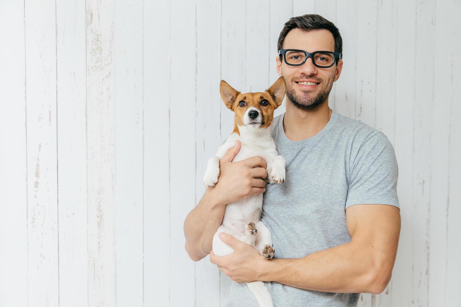 atractivo y alegre hombre caucásico con camiseta informal, espectáculos, tiene una mascota favorita, se alza contra un fondo blanco de madera con un espacio en blanco. hombre feliz sin afeitar con su jack russell terrier foto