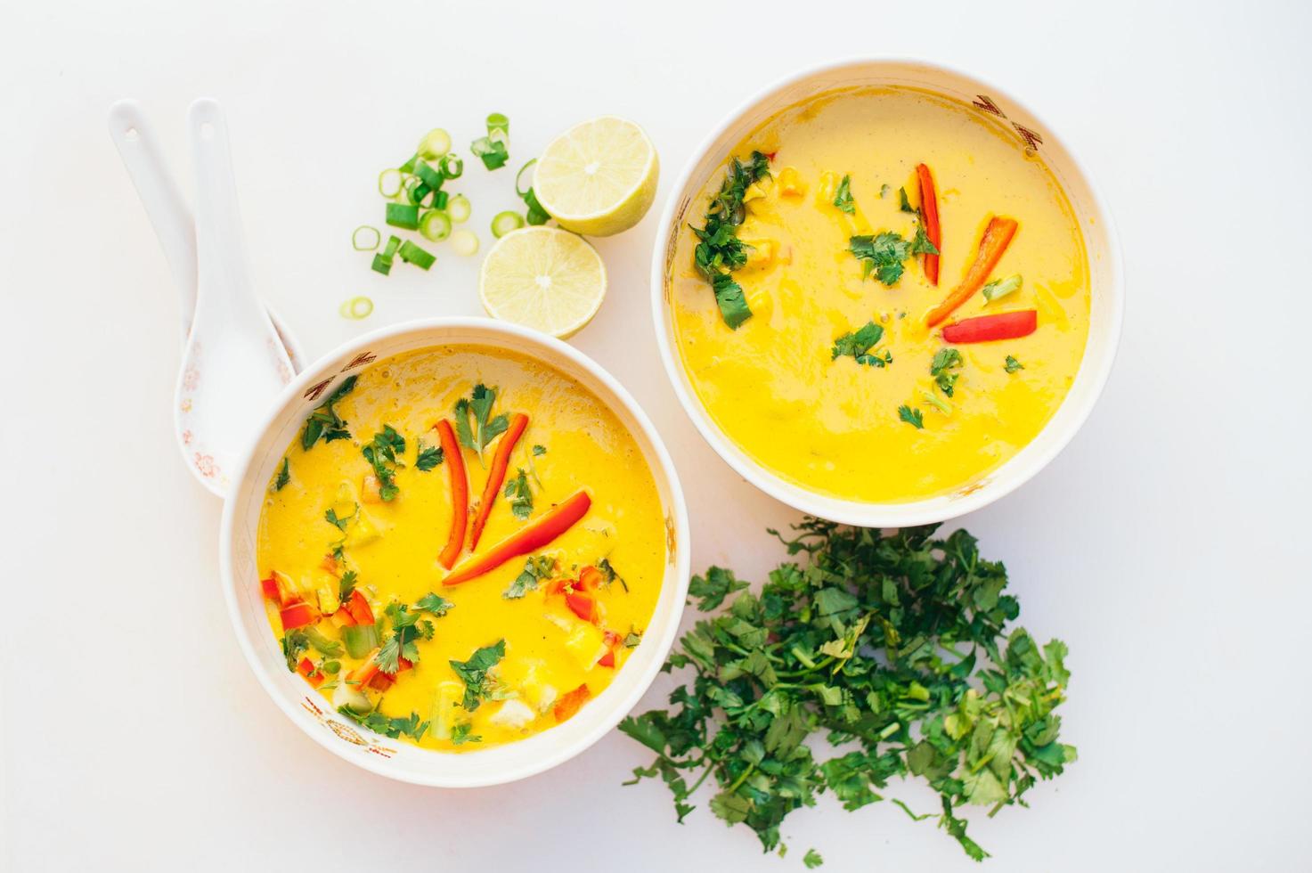 Two bowls of fresh coconut curry soup with cut green parsley, slices of lime, spoons, isolated over white background. Curry bowl for dinner. Vegetarian food concept photo