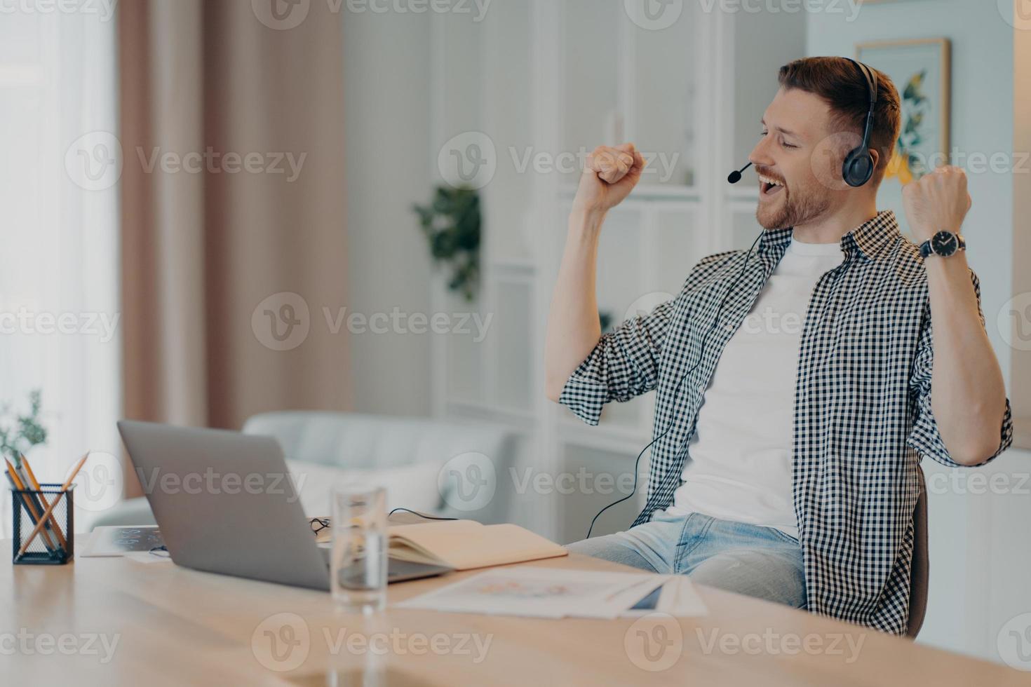 feliz hombre exitoso aprieta los puños celebra el triunfo trabaja en línea usa poses de auriculares en el escritorio con una computadora portátil vestida de manera informal. un estudiante varón muy contento escucha una conferencia educativa en línea foto