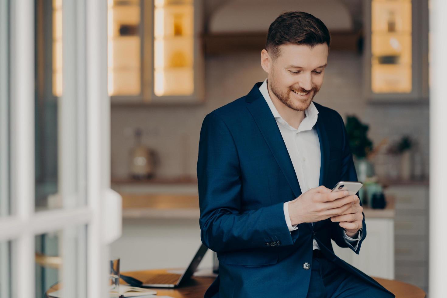 Business lifestyle. Glad handsome man with stubble uses mobile phone for texting online checks newsfeed wears elegant clothes posesagainst blurred coworking space. Focused salesman with gadget photo