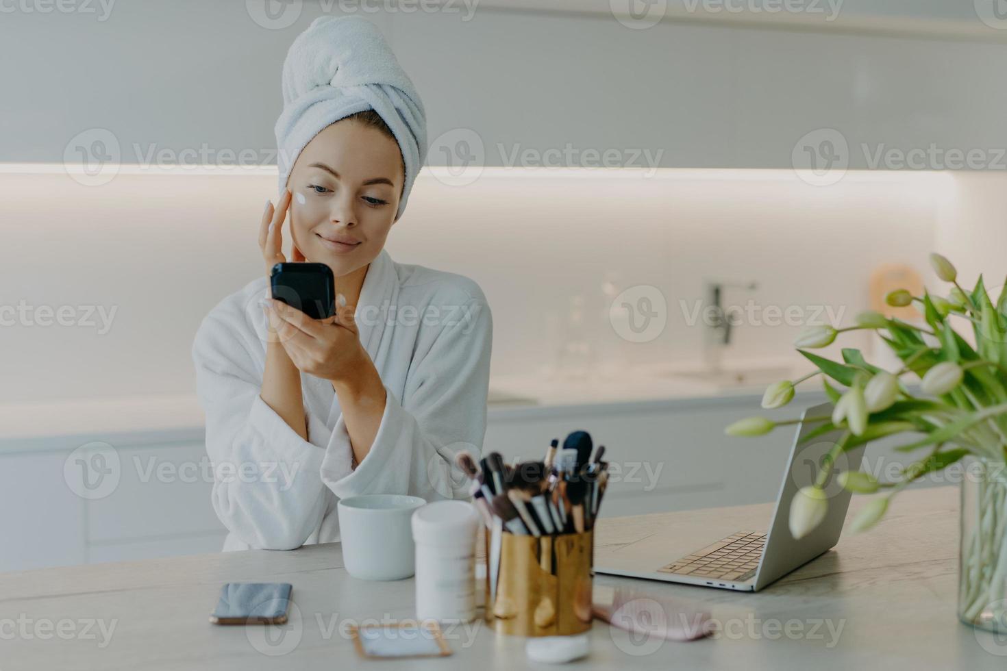 foto de una mujer de buen aspecto que se aplica crema facial en la cara se mira en el espejo se preocupa por la tez usa una toalla de baño y una bata se sienta en la mesa contra el interior de la cocina. concepto de tiempo de belleza