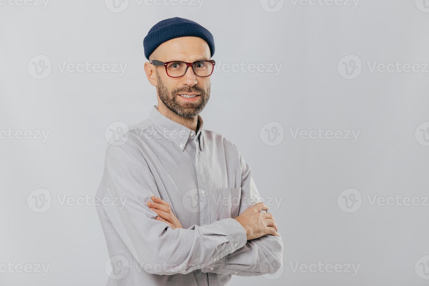 Isolated shot of pleasant looking unshaven male keeps hands crossed, wears stylish hat, optical glasses, models over white background with copy space for your advertising content, enjoys leisure photo