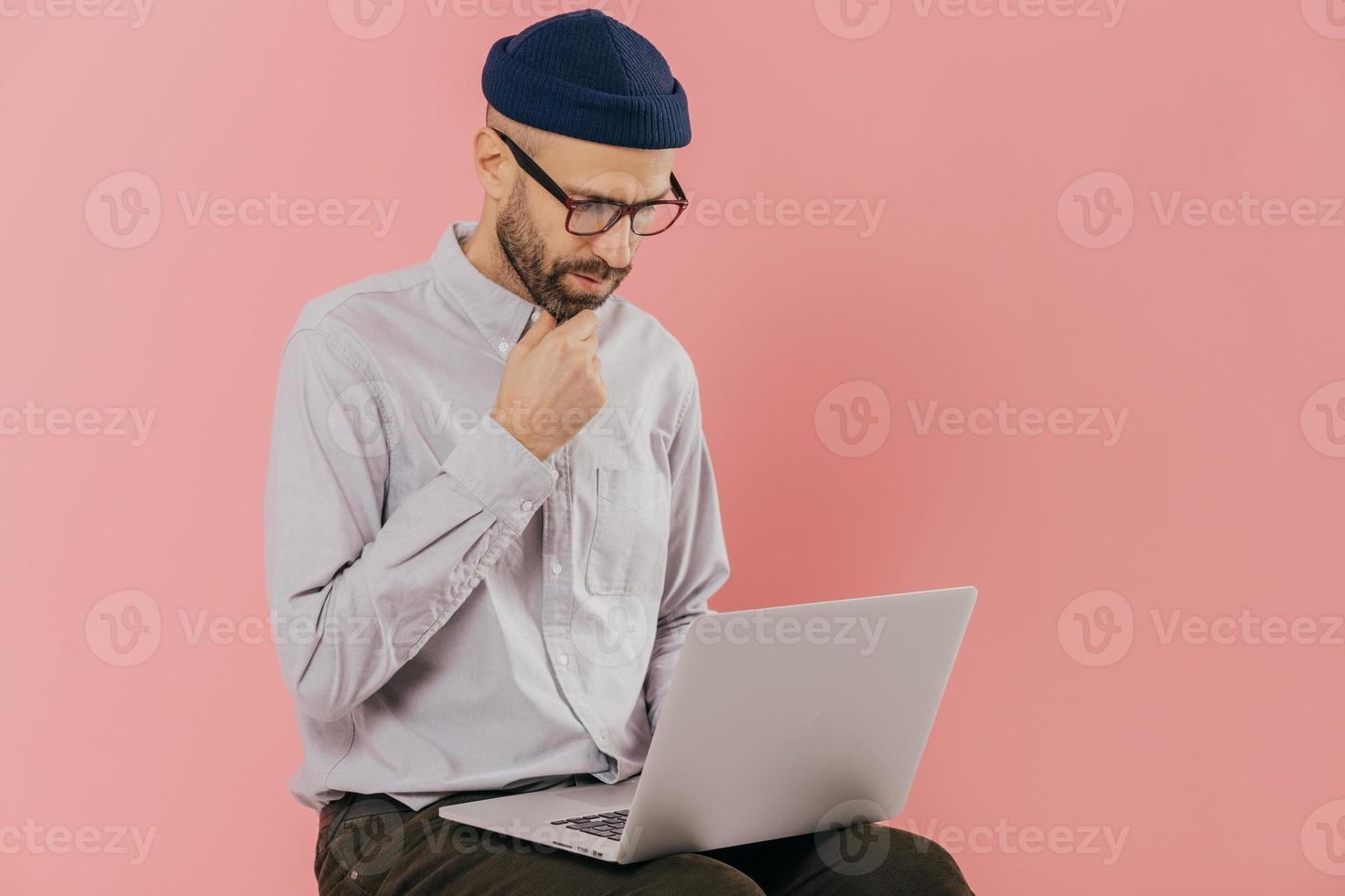 Photo of concentrated unshaven man holds chin, looks attentively at screen of laptop computer, enjoys online communication, wears optical glasses and formal outfit, isolated over pink background