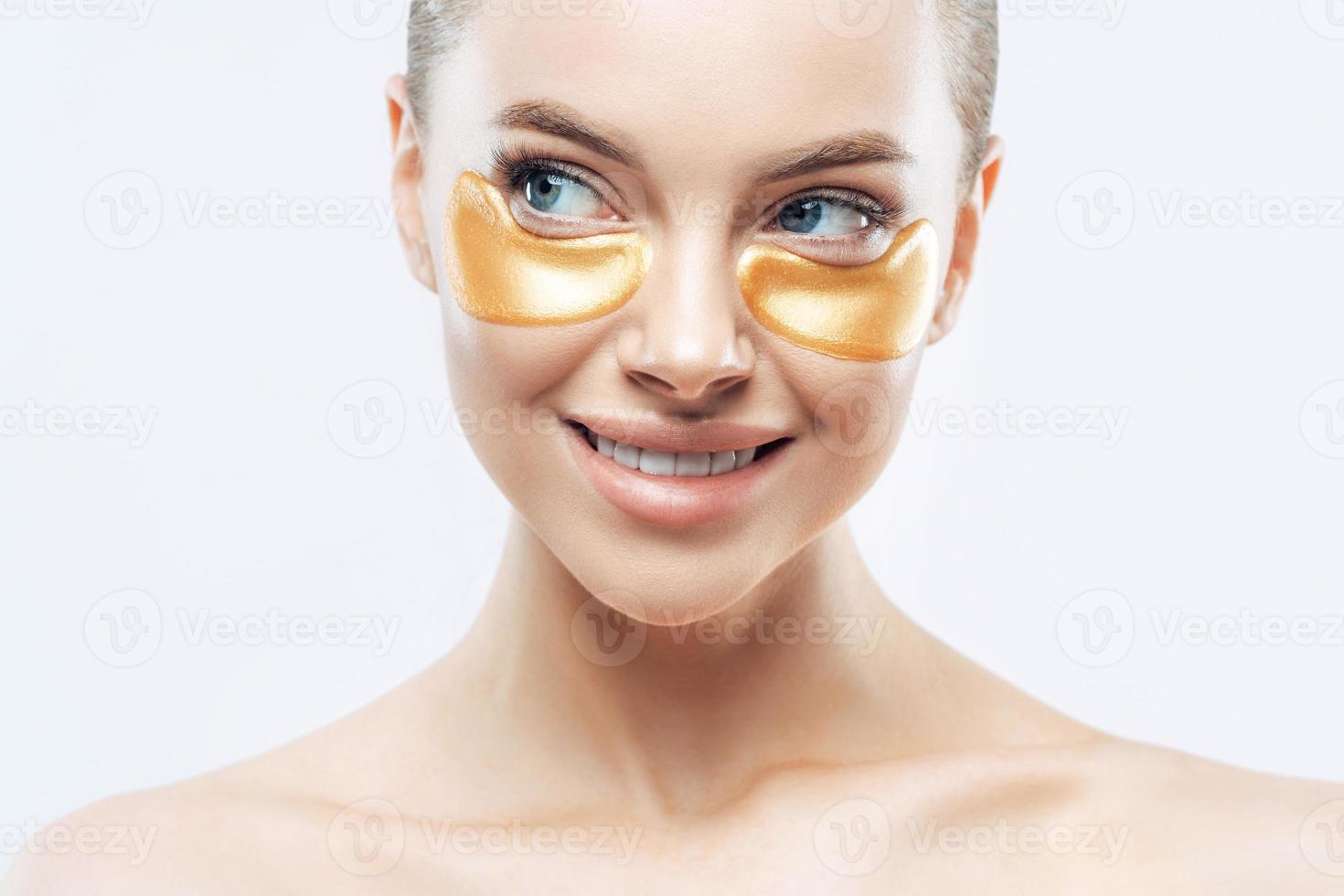 Cropped image of beautiful woman with naked shoulders, performs daily face care routine, applies golden patches under eyes to remove dark circles, isolated on white background. Beauty concept photo