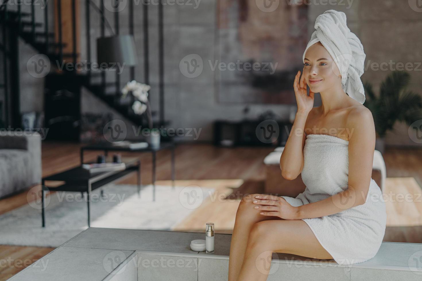 Satisfied female model applies cosmetic product for healthy skin after showering, uses moisturizer and has daily routine, wrapped in bath towel, poses against home interior. Skin care procedure photo