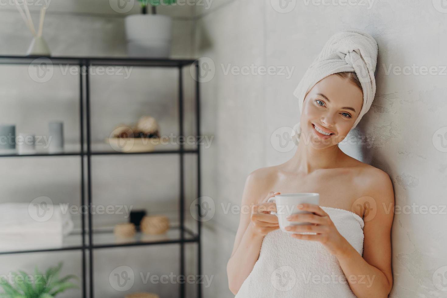 una mujer muy sonriente con una piel sana envuelta en una toalla blanca, se alegra en el interior, bebe café, tiene una expresión facial positiva, posa en el interior. personas, spa, mimos, cuidado de la piel y concepto de bienestar foto