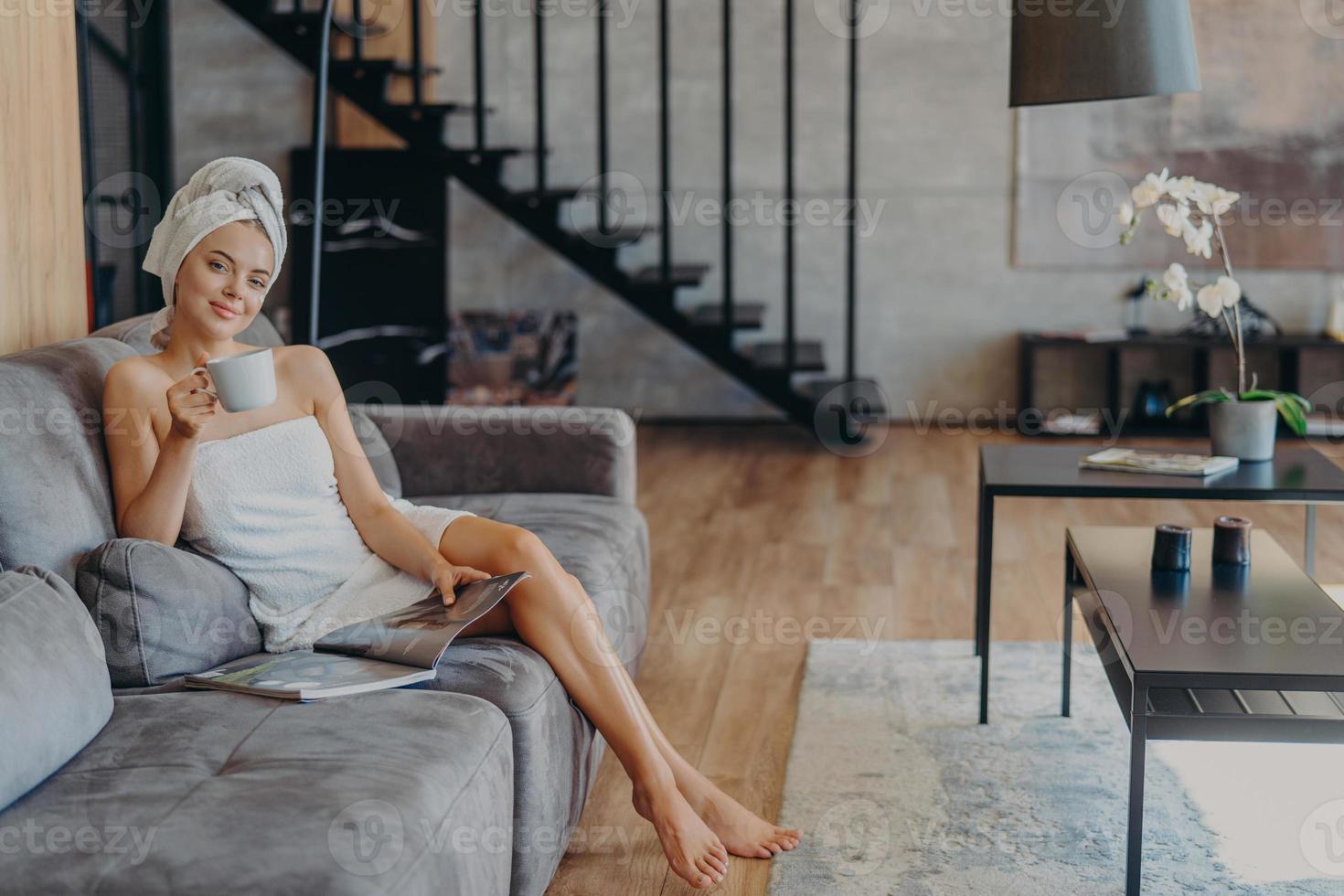 Happy young woman poses wrapped in bath towel, drinks aromatic coffee and reads magazine, sits on comfortable sofa, poses in living room at home. Domestic atmoshpere, leisure time, lifestyle photo