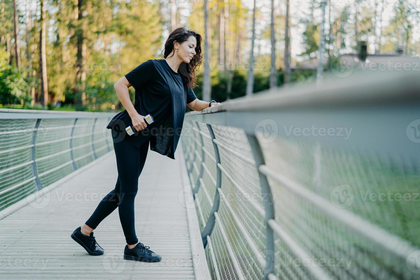 Active brunette woman dressed in casual t shirt and leggings, holds dumbbells, poses at bridge, looks down, goes in for sport outdoor, leads healthy lifestyle, lifts weight, uses sport equipment photo