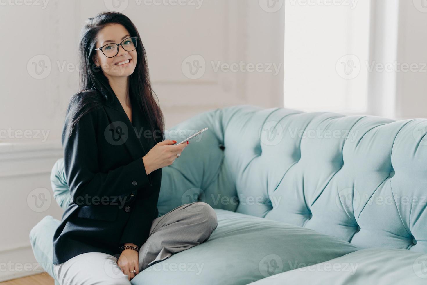 Happy business lady in official clothes, sits on comfortable sofa, uses mobile phone, reads news via network, searches information in cellular, going to phone partner, explores online project photo