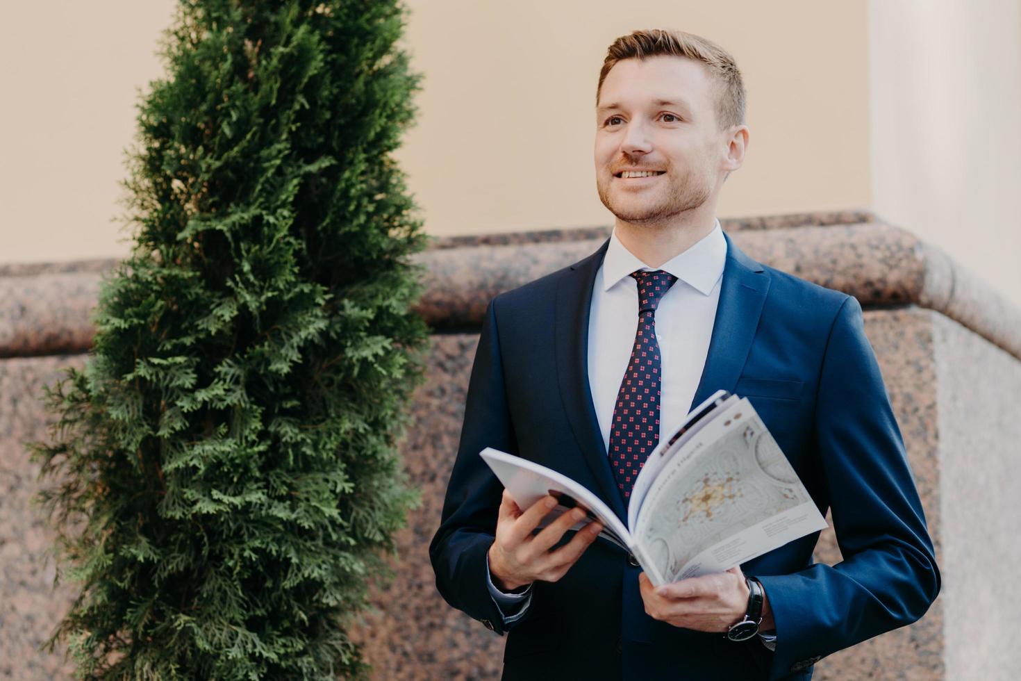 Horizontal shot of attractive male with joyful expression, being focused into distance, has stubble, dressed in formal clothes, holds city map, tries to orientate in big unknown city has smile on face photo