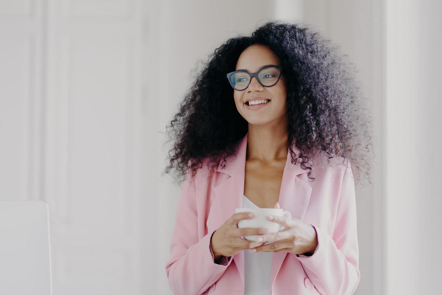 foto de una próspera empresaria afroamericana de buen aspecto que espera a su socio en el cargo, bebe café y discute planes futuros con un colega que mira hacia otro lado con expresión alegre y viste ropa formal