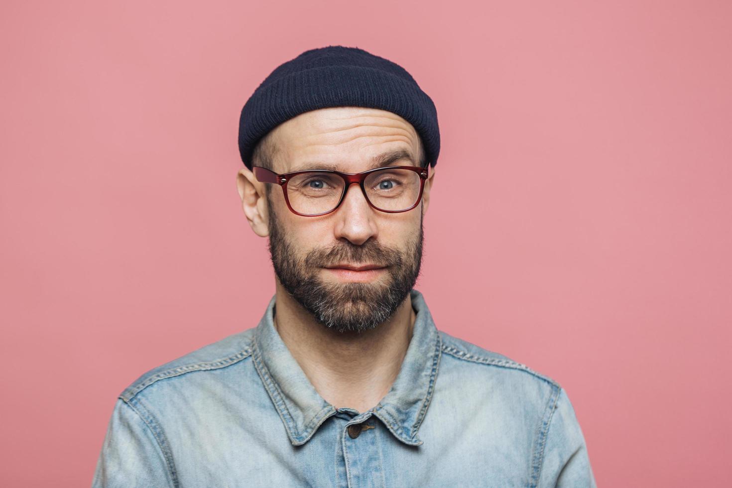 Portrait of handsome bearded man looks with pleased surprised expression, wears glasses and black hat, isolated over pink studio background. Middle aged unshaven male expresses bewilderment. photo