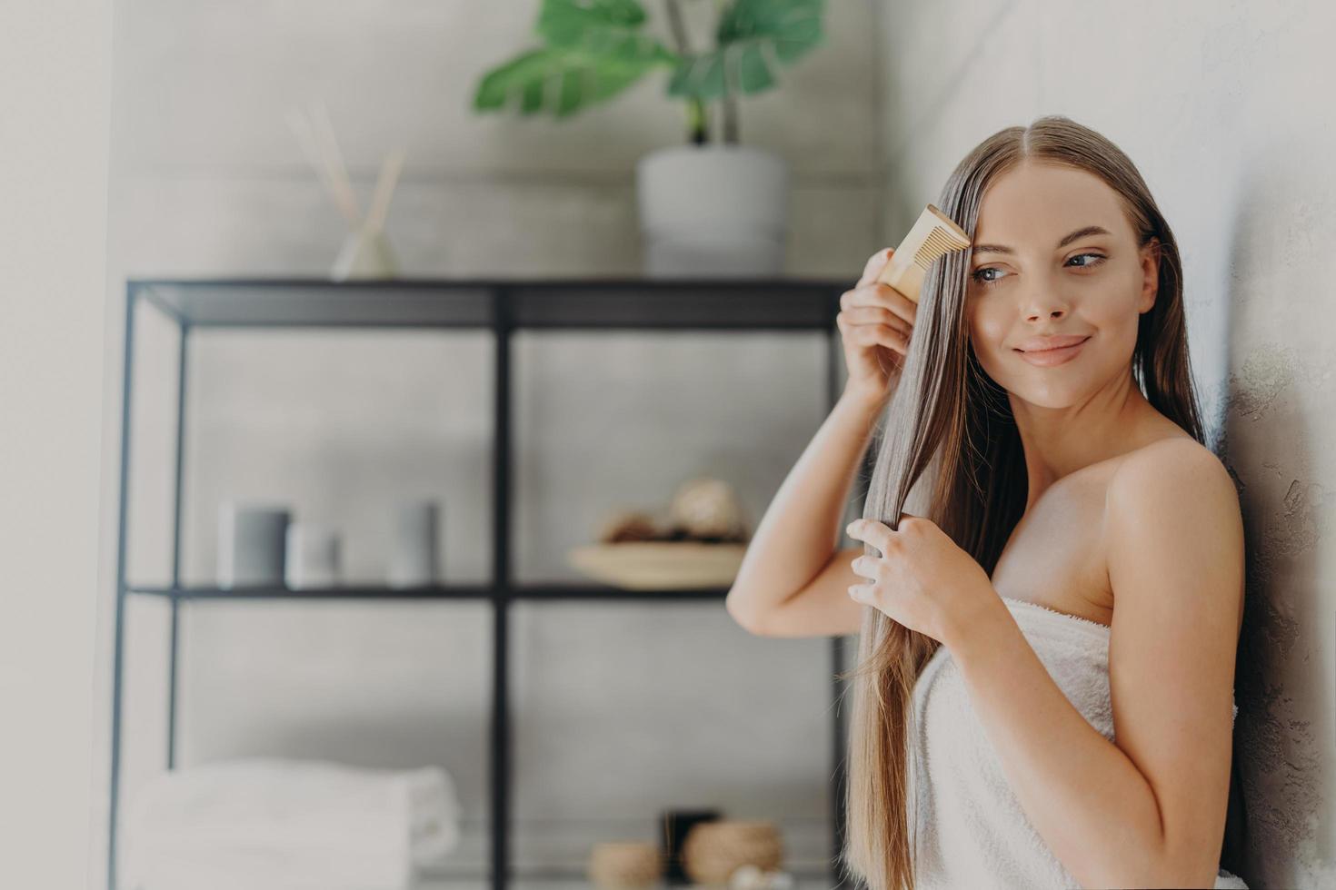 atractiva mujer joven sonríe y disfruta de su belleza, peina el pelo largo y recto después de aplicar la máscara, se para envuelta en una toalla de baño en un baño acogedor, tiene la piel suave. concepto de cuidado del cabello, spa y belleza foto