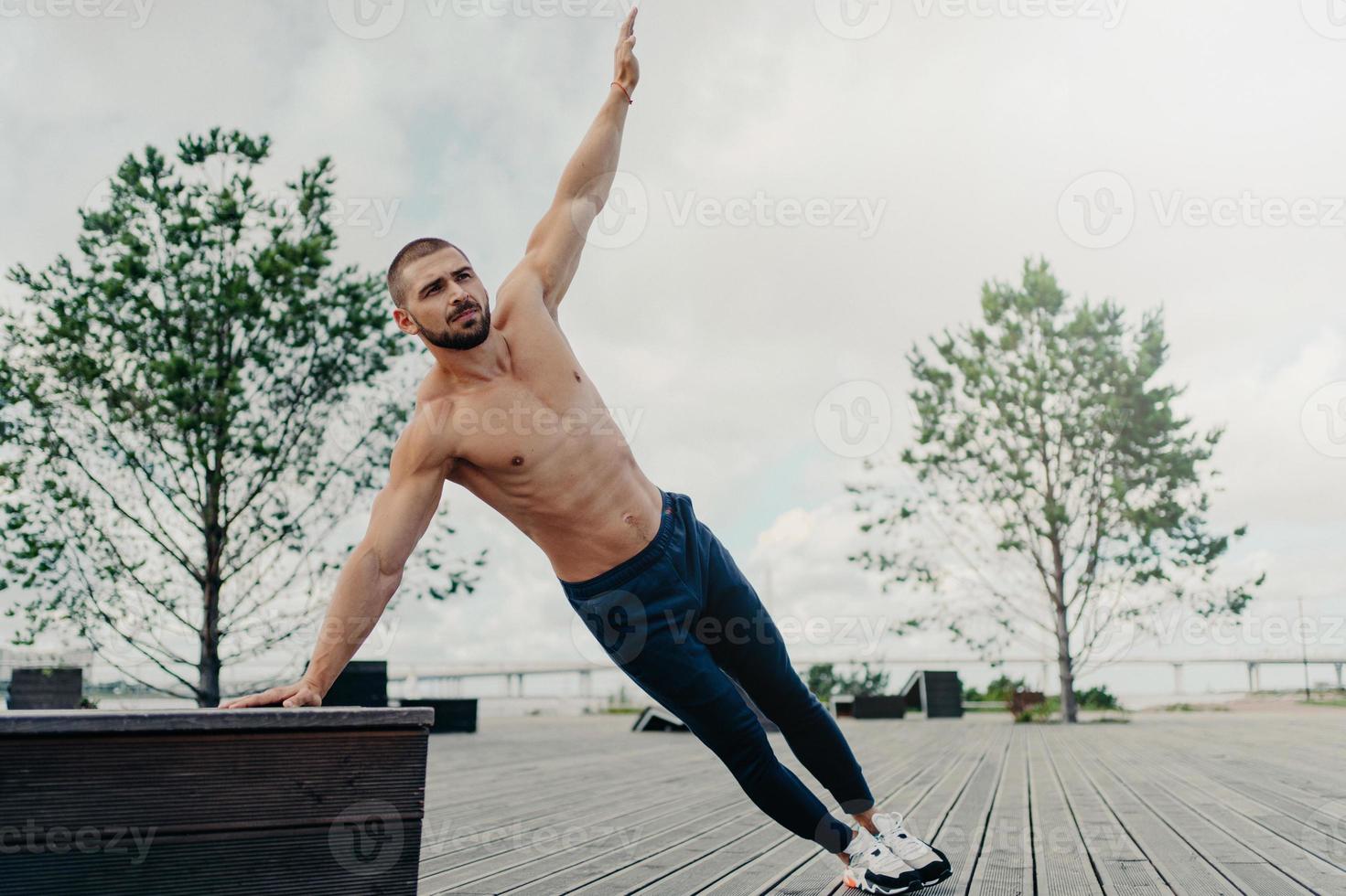Fit healthy muscular man stands in side plank and raises arm, poses with naked torso, dressed in sport trousers and sneakers, exercises abs, raises abdominal crunch, trains outdoor near beach photo